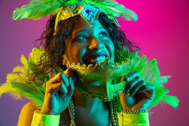 Soñadora. Hermosa mujer joven en carnaval, elegante disfraz de mascarada con plumas bailando en la pared degradada en neón. Concepto de celebración navideña, tiempo festivo, baile, fiesta, diversión.