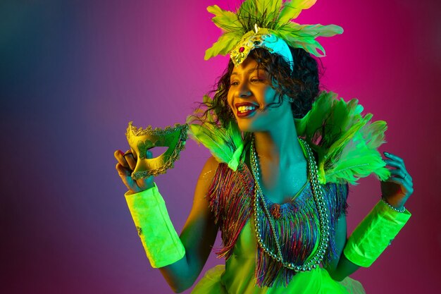 Soñadora. Hermosa mujer joven en carnaval, elegante disfraz de mascarada con plumas bailando sobre fondo degradado en neón. Concepto de celebración navideña, tiempo festivo, baile, fiesta, diversión.