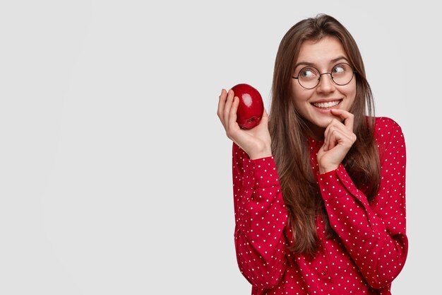 Soñadora joven europea mantiene el dedo cerca de los labios, sostiene una deliciosa manzana fresca, piensa en cocinar un pastel