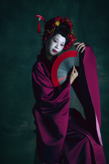 Soñadora. Joven japonesa como geisha aislada en la pared de color verde oscuro. Estilo retro, comparación del concepto de eras. Modelo femenino hermoso como personaje histórico brillante, anticuado.