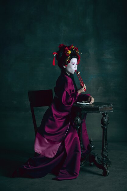 Soñadora. Joven japonesa como geisha aislada en la pared de color verde oscuro. Estilo retro, comparación del concepto de eras. Modelo femenino hermoso como personaje histórico brillante, anticuado.