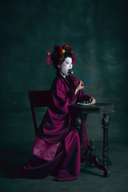 Soñadora. Joven japonesa como geisha aislada en la pared de color verde oscuro. Estilo retro, comparación del concepto de eras. Modelo femenino hermoso como personaje histórico brillante, anticuado.