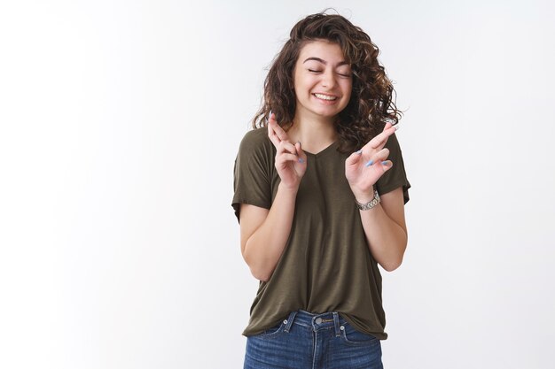 Soñadora linda chica joven afortunada rezando creer milagro cerrar ojos sonriendo ampliamente cruzar los dedos mientras hace un deseo pidiendo a Dios con suerte cumplir el sueño de pie fondo blanco anticipando fielmente