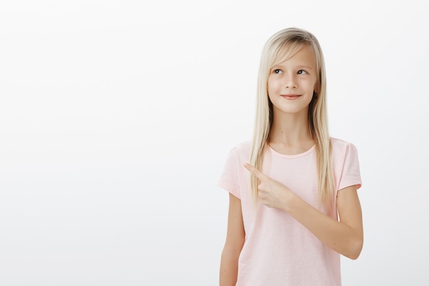 Soñadora linda niña rubia apuntando hacia la esquina superior izquierda complacida, imaginando o pensando