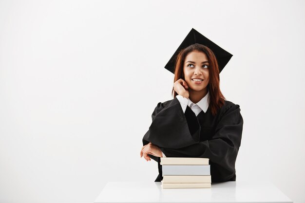 Soñadora mujer africana graduada sonriendo pensando sentado con libros.