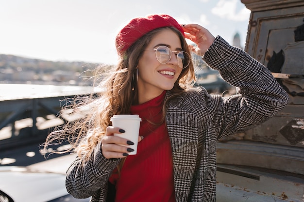 Soñadora mujer francesa de pelo largo con gafas mirando a otro lado con una sonrisa, sosteniendo una taza de café