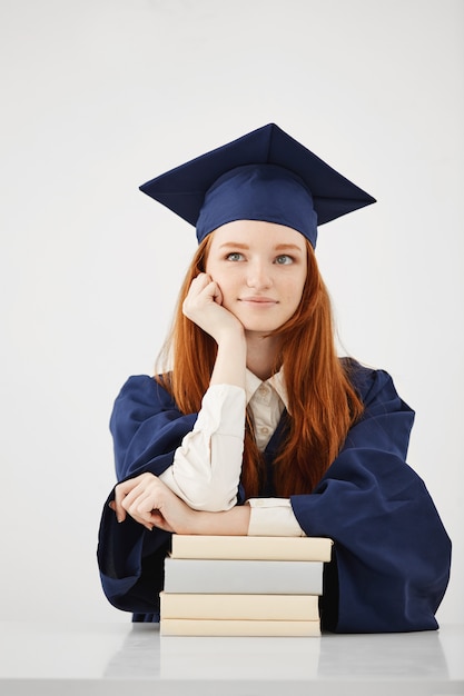 Soñadora mujer graduada sonriendo pensando sentado con libros sobre superficie blanca