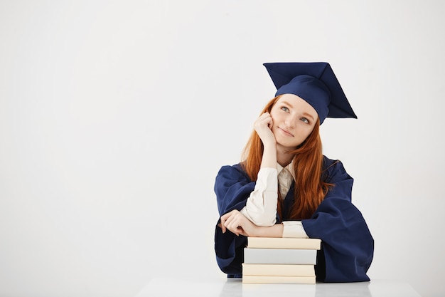 Soñadora mujer graduada sonriendo pensando sentado con libros.