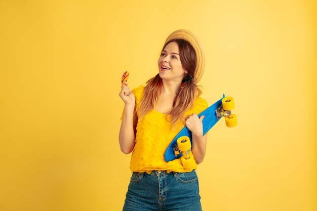 Soñadora, sostiene skate. Retrato de mujer caucásica sobre fondo amarillo de estudio.