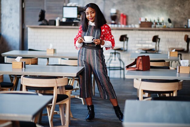 Sonfident joven mujer afroamericana en ropa casual elegante en el café con una taza de bebida caliente en la mano