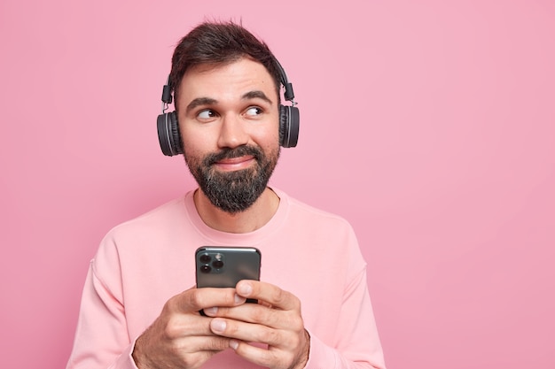 Sonido encendido. Hombre barbudo guapo complacido escucha música a través de auriculares desde la lista de reproducción sostiene el teléfono móvil usa una nueva aplicación mira hacia otro lado pensativamente