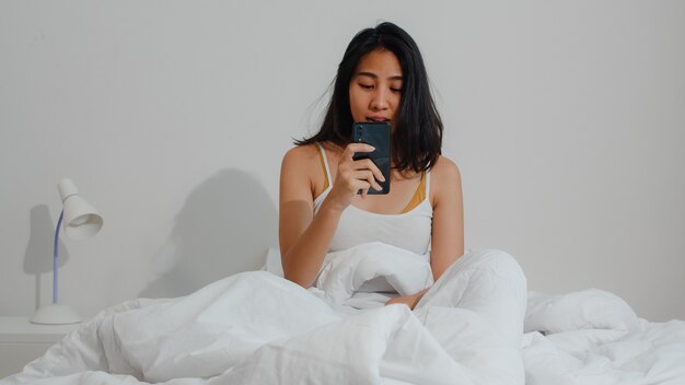 Soñolienta joven asiática sorprendida cuando se levanta tarde para una cita en la cama en la habitación de su casa en la mañana. La mujer india no sonó la alarma del teléfono móvil y se ha quedado dormida.