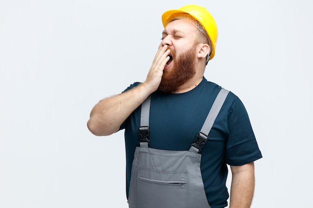 Soñoliento joven trabajador de la construcción con casco de seguridad y uniforme manteniendo la mano en la boca bostezando con los ojos cerrados aislado sobre fondo blanco.