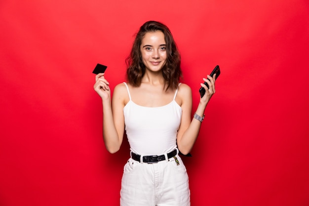 Sonríe felizmente a la mujer que sostiene el teléfono inteligente y la tarjeta de crédito de compras en línea en la pared roja.