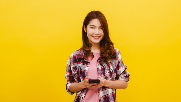 Sonriendo adorable mujer asiática con teléfono con expresión positiva, sonríe ampliamente, vestido con ropa casual y mirando a la cámara sobre la pared amarilla. Feliz adorable mujer alegre disfruta el éxito.