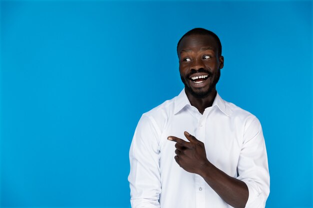 Sonriendo afroamericano barbudo en camisa blanca sobre fondo azul está mostrando algo