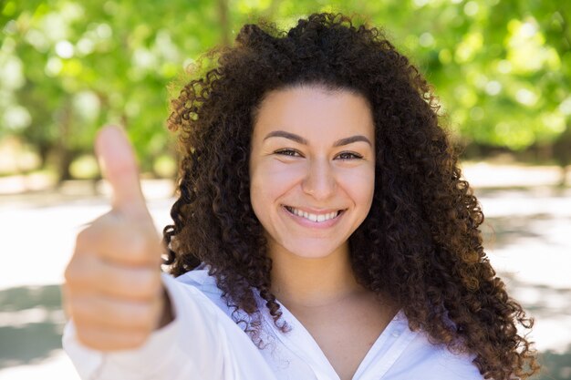 Sonriendo bastante dama de pelo rizado mostrando el pulgar hacia arriba en el parque