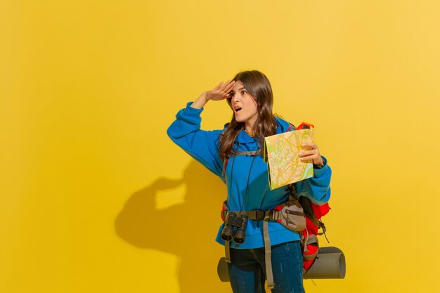 Sonriendo, buscando el camino. Retrato de una alegre joven turista caucásica con bolsa y binoculares aislado sobre fondo amarillo de estudio. Preparándose para viajar. Resort, emociones humanas, vacaciones.