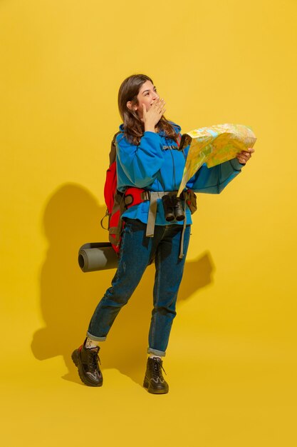 Sonriendo, buscando el camino. Retrato de una joven turista caucásica alegre con bolsa y binoculares aislado sobre fondo amarillo de estudio.