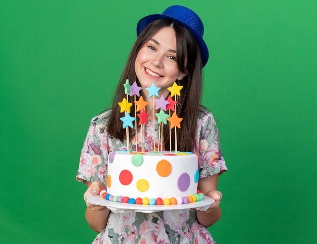 Sonriendo la cabeza inclinada joven hermosa chica con sombrero de fiesta sosteniendo pastel aislado en la pared verde