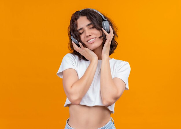 Sonriendo complacida joven hermosa mujer usando audífonos y poniendo las manos sobre ellos con los ojos cerrados en la pared naranja aislada con espacio de copia