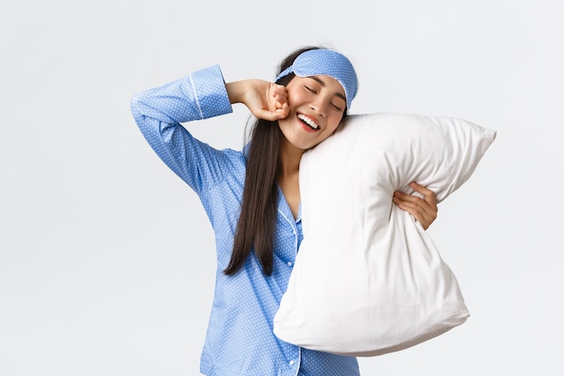 Sonriendo complacida linda chica asiática en pijama azul y máscara para dormir, abrazando la almohada y estirando las manos encantadas cuando finalmente se va a la cama, quiere dormir o despertarse en la mañana, fondo blanco.