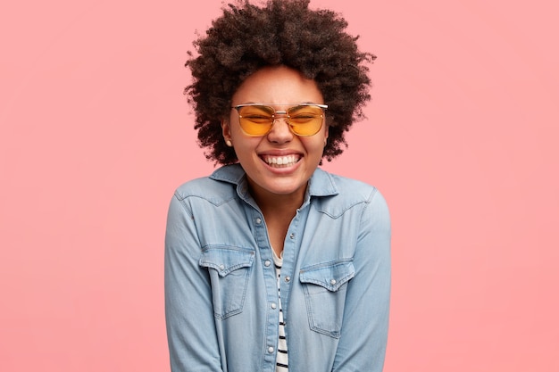 Sonriendo contenido niña de piel oscura cierra los ojos y sonríe ampliamente, está deprimido, se ríe de una broma divertida, usa camisa vaquera, modelos sobre una pared rosa. Buen concepto de emociones y sentimientos.