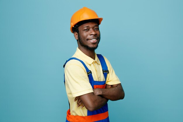 Sonriendo cruzando las manos joven constructor afroamericano en uniforme aislado sobre fondo azul.