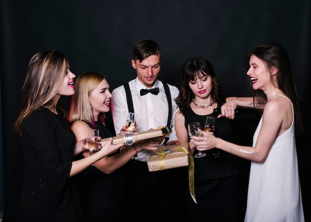 Sonriendo damas y chicos en ropa de noche con vasos de bebidas y cajas de regalo.