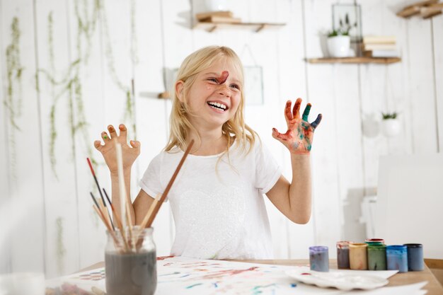 Sonriendo con dientes linda rubia pequeña mostrando sus manos en pintura. Alegre niña de siete años ocupada con dibujo de pintura sin desorden.