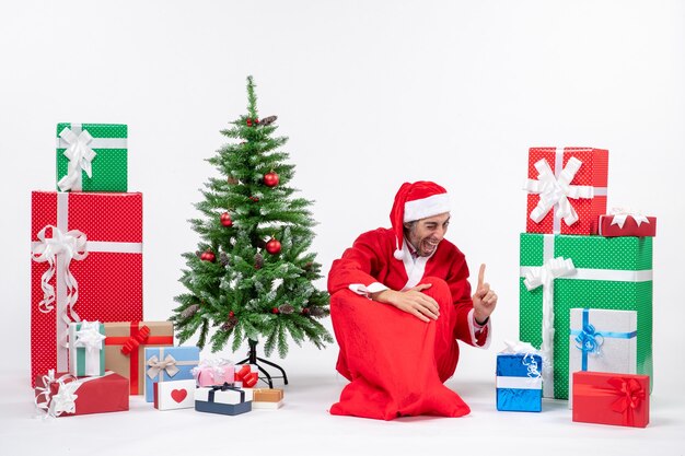 Sonriendo divertido emocionado joven vestido como Papá Noel con regalos y árbol de Navidad decorado sentado en el suelo apuntando arriba sobre fondo blanco.