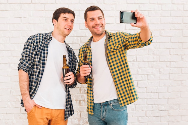Sonriendo dos amigos varones sosteniendo una botella de cerveza tomando selfie en el teléfono móvil