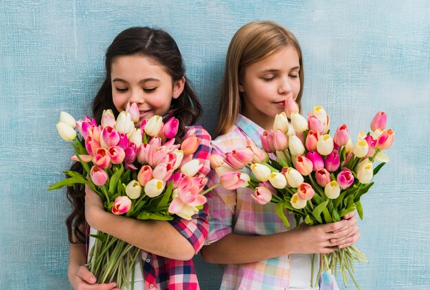 Sonriendo dos chicas de pie contra la pared azul que huele la flor de tulipanes