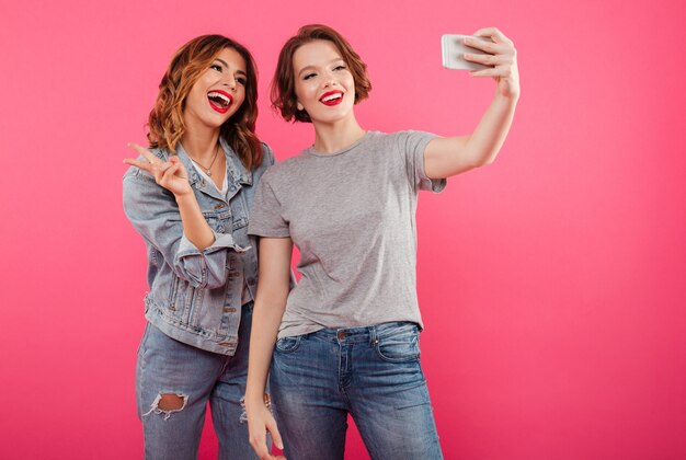 Sonriendo dos mujeres emocionales hacen selfie por teléfono.