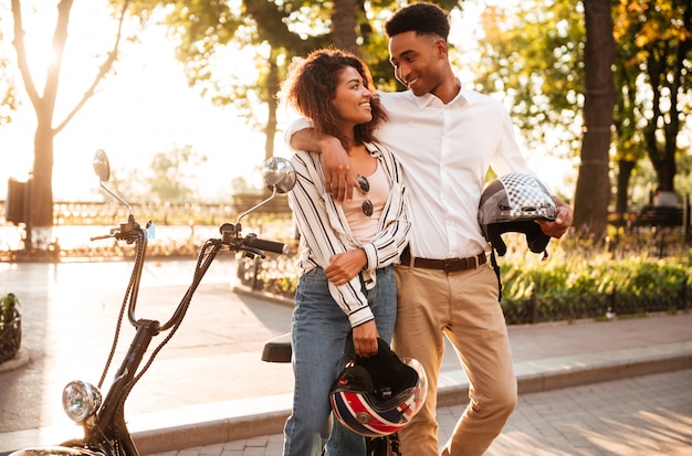 Sonriendo ee pareja africana abrazando cerca de la moto moderna en el parque