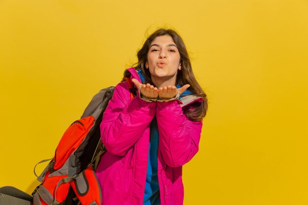 Sonriendo, enviando un beso. Retrato de una joven turista caucásica alegre con bolsa y binoculares aislado sobre fondo amarillo de estudio.
