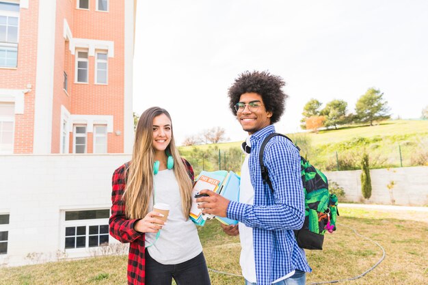 Sonriendo estudiantes jóvenes masculinos y femeninos que sostienen la taza y los libros de café para llevar en la mano que se coloca en el campus