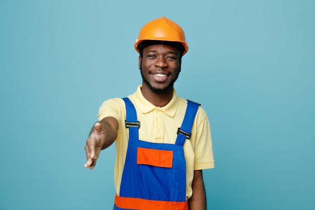 Sonriendo extendiendo la mano a la cámara joven constructor afroamericano en uniforme aislado sobre fondo azul.