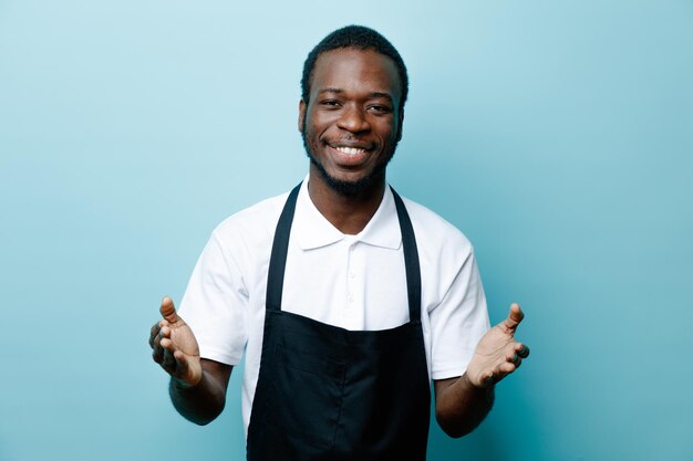 Sonriendo extendiendo las manos joven peluquero afroamericano en uniforme aislado sobre fondo azul.