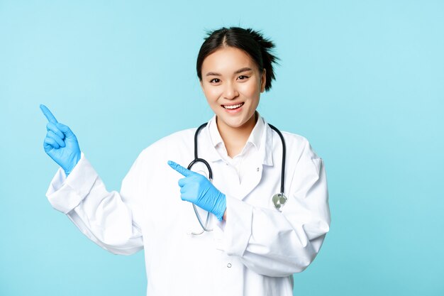 Sonriendo feliz doctora o enfermera, señalando con el dedo hacia la izquierda, con uniforme médico y guantes, mostrando publicidad del hospital, fondo azul