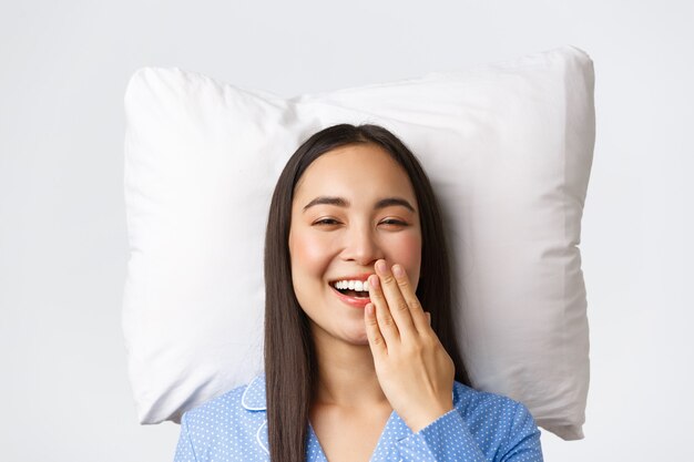 Sonriendo feliz hermosa niña asiática acostada en la cama sobre la almohada en pijama azul, despertando los ojos abiertos y bostezando, rutina matutina de mujer joven. Linda hembra en pijamas quedarse en cama, fondo blanco.