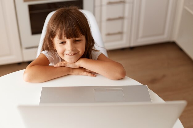 Sonriendo feliz linda niña de cabello oscuro sentado en la mesa, mirando la pantalla del portátil, viendo dibujos animados interesantes, posando en la sala de luz en casa.