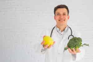 Foto gratuita sonriendo feliz médico masculino ofreciendo manzana y brócoli