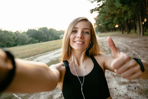 Sonriendo a hermosa chica en ropa deportiva tomando un selfie, mostrando un gran dedo
