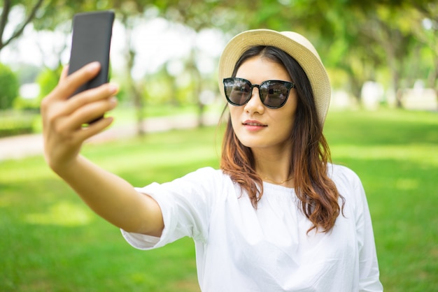 Sonriendo a hermosa mujer asiática en gafas de sol tomando selfie en el parque de verano.