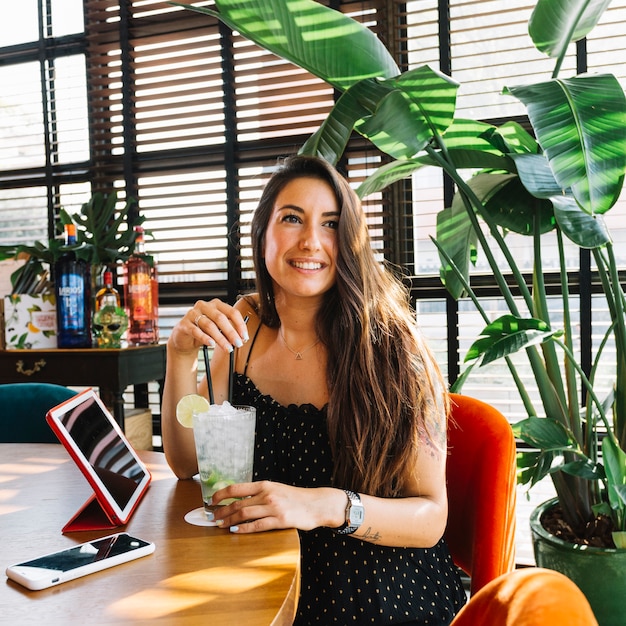 Sonriendo hermosa mujer joven con cóctel; Smartphone y tableta digital en el restaurante.