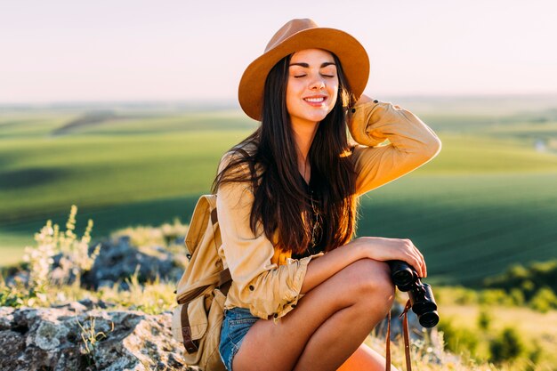 Sonriendo a hermosa mujer sentada en roca sosteniendo binoculares