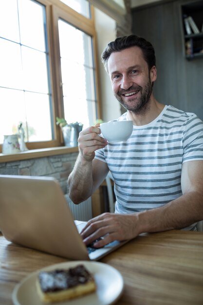 Sonriendo el hombre usando la computadora portátil y que tiene una taza de café en la cafetería