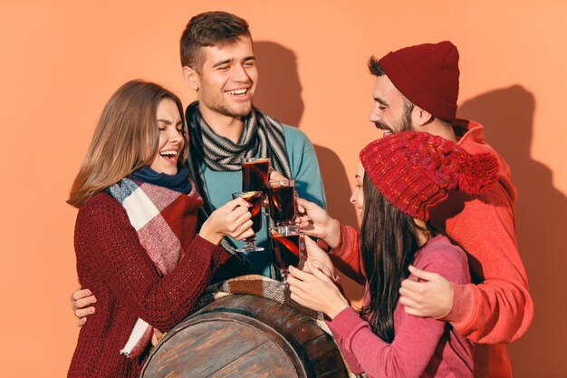 Sonriendo a hombres y mujeres europeos durante la sesión de fotos del partido. Los chicos se hacen pasar por amigos en el festival de estudio con copas con vino caliente caliente en primer plano.