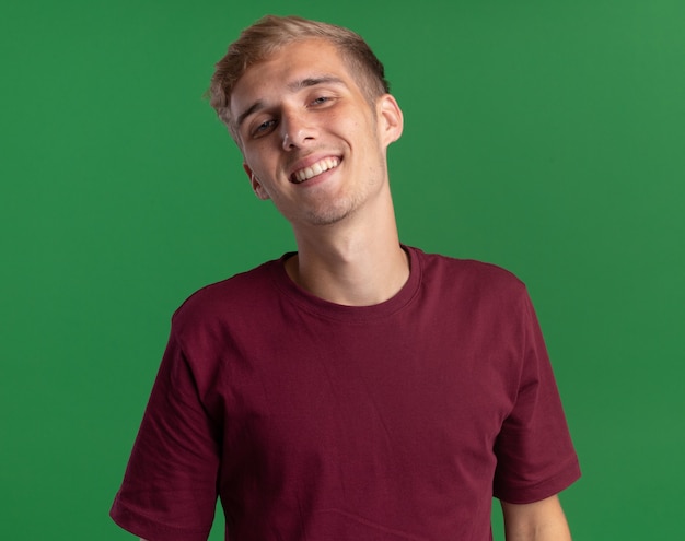 Sonriendo inclinando la cabeza joven guapo con camisa roja aislado en la pared verde